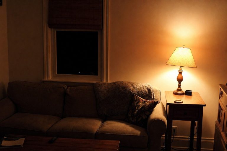 An ordinary living-room corner at night lit only by a warm table lamp casting a soft orange pool of light on a plain sofa and side table